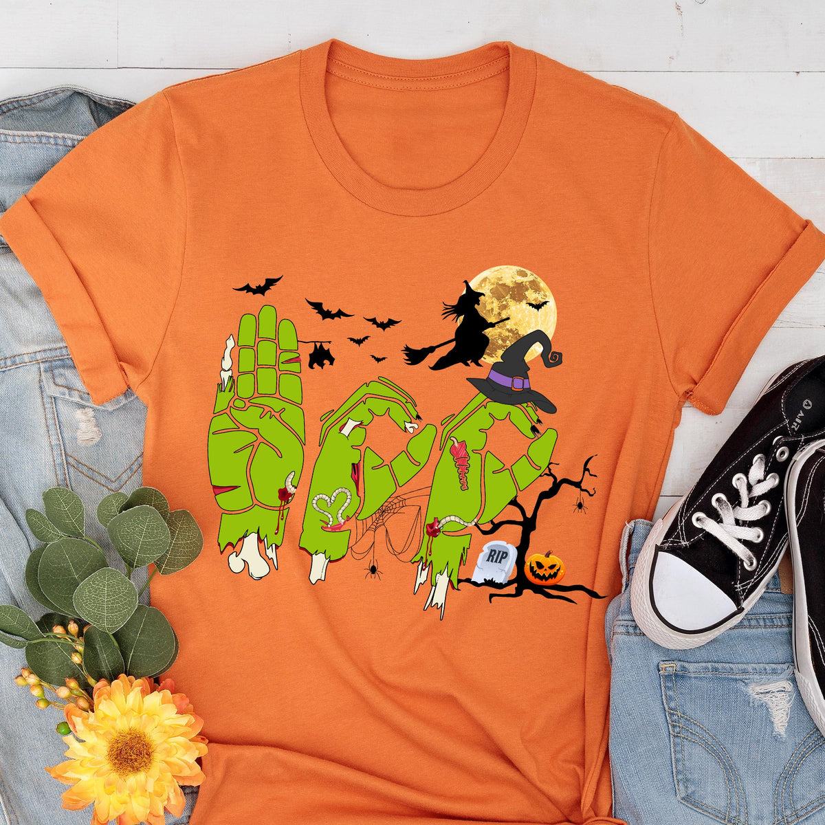 May include: An orange short-sleeve t-shirt featuring a graphic of three green zombie hands forming the word BOO in American Sign Language. The design includes a witch on a broomstick, a full moon, bats, a spider web, a tombstone, and a jack-o'-lantern.