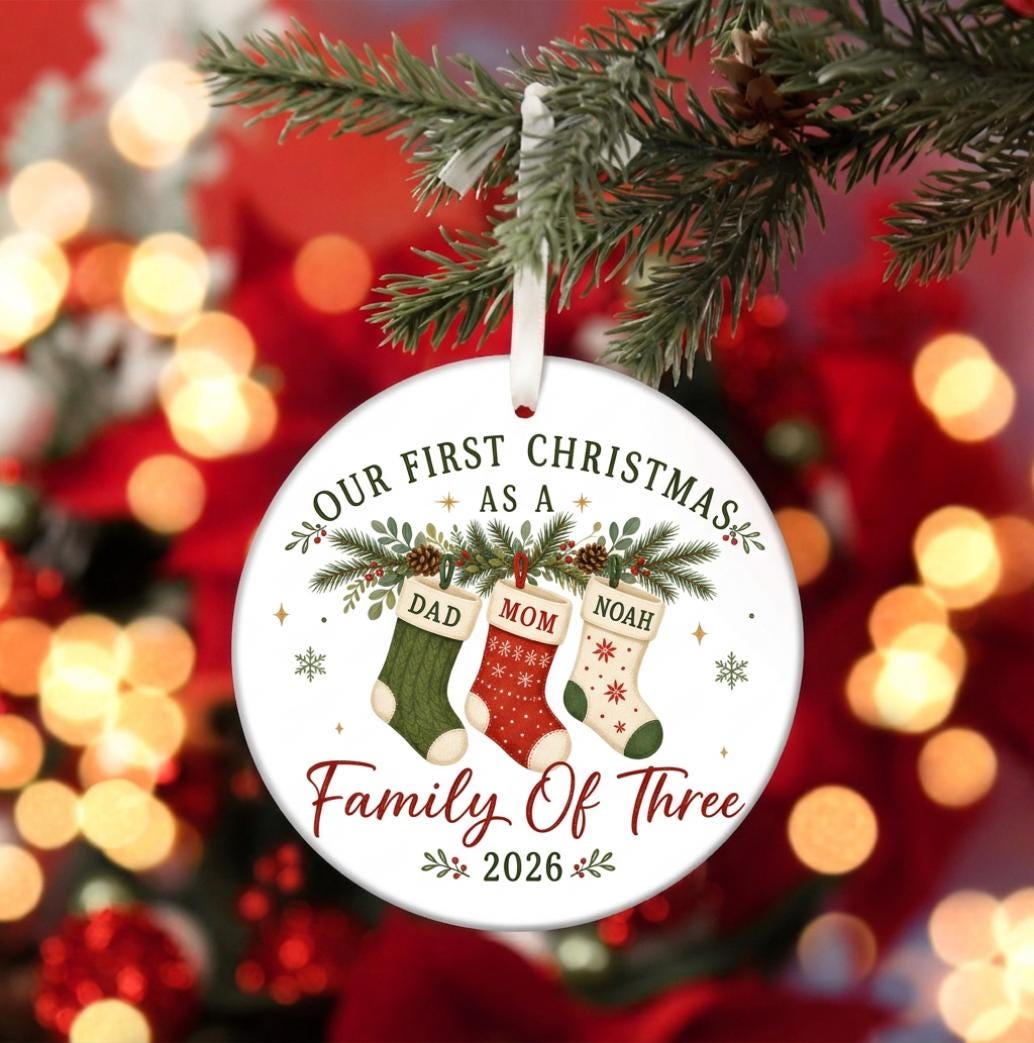 May include: A round white ceramic Christmas ornament hanging from a white ribbon. The design features three stockings labeled DAD, MOM, and NOAH, with the text OUR FIRST CHRISTMAS AS A FAMILY OF THREE and the year 2026 printed in red and black.