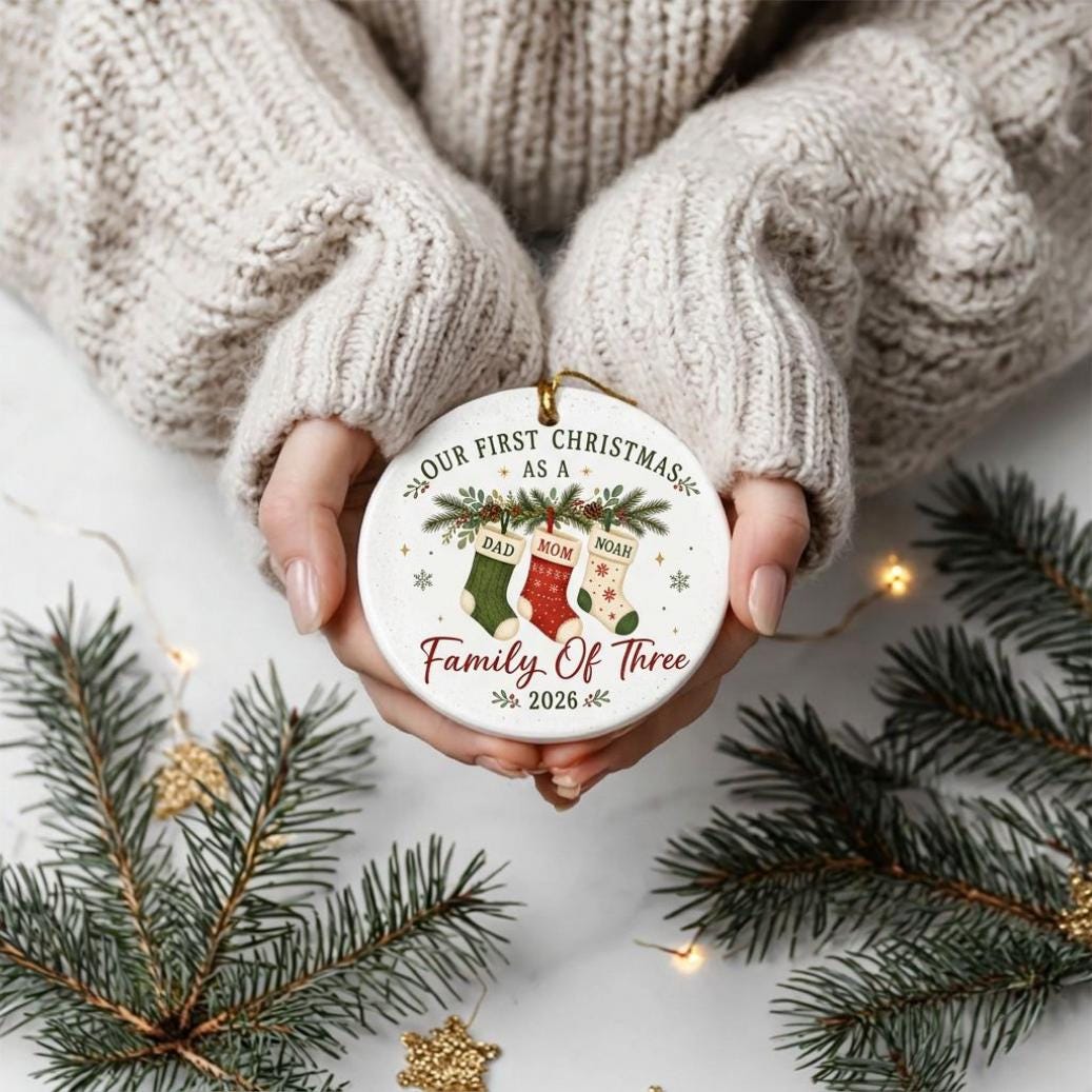 May include: A round white ceramic ornament featuring a festive design with three hanging stockings labeled Dad, Mom, and Noah. The text reads Our First Christmas as a Family of Three 2026, accented with green pine branches and small snowflakes.
