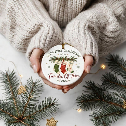 May include: A round white ceramic ornament featuring a festive design with three hanging stockings labeled Dad, Mom, and Noah. The text reads Our First Christmas as a Family of Three 2026, accented with green pine branches and small snowflakes.