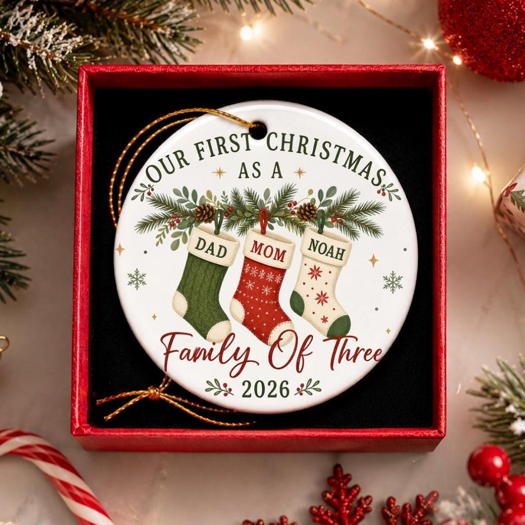 May include: A round white ceramic Christmas ornament featuring the text "OUR FIRST CHRISTMAS AS A Family Of Three 2026" and an illustration of three hanging stockings labeled "DAD," "MOM," and "NOAH" decorated with pine branches and pinecones.