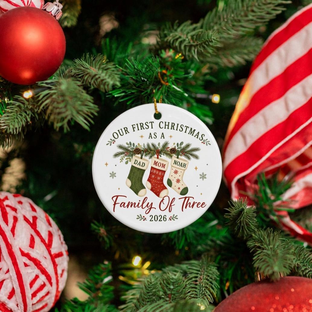 May include: A round white ceramic ornament featuring the text Our First Christmas As A Family Of Three 2026. The design includes three hanging Christmas stockings labeled Dad, Mom, and Noah, decorated with pine branches and small snowflakes.