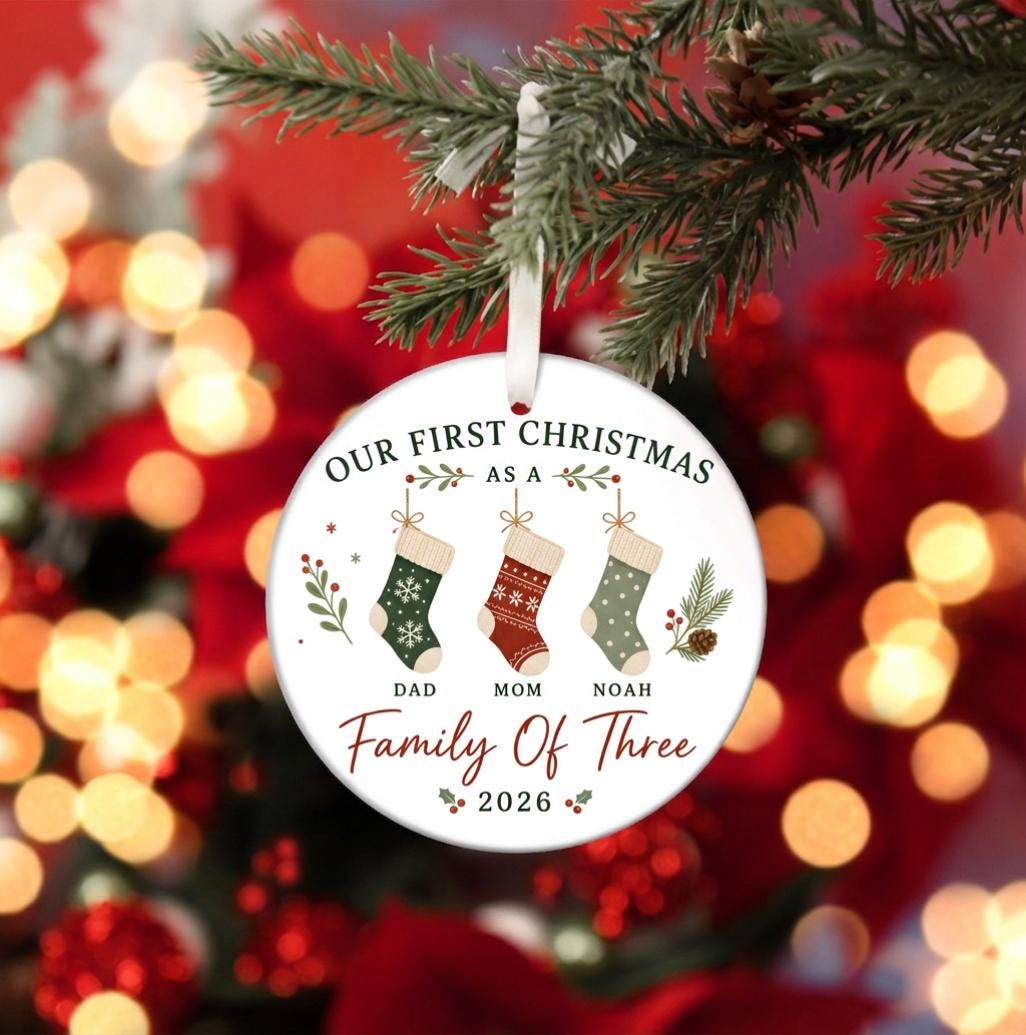 May include: A round white ceramic Christmas ornament hanging from a white ribbon. It features the text "OUR FIRST CHRISTMAS AS A Family Of Three" and "2026" with illustrations of three stockings labeled "DAD", "MOM", and "NOAH" with green and red accents.