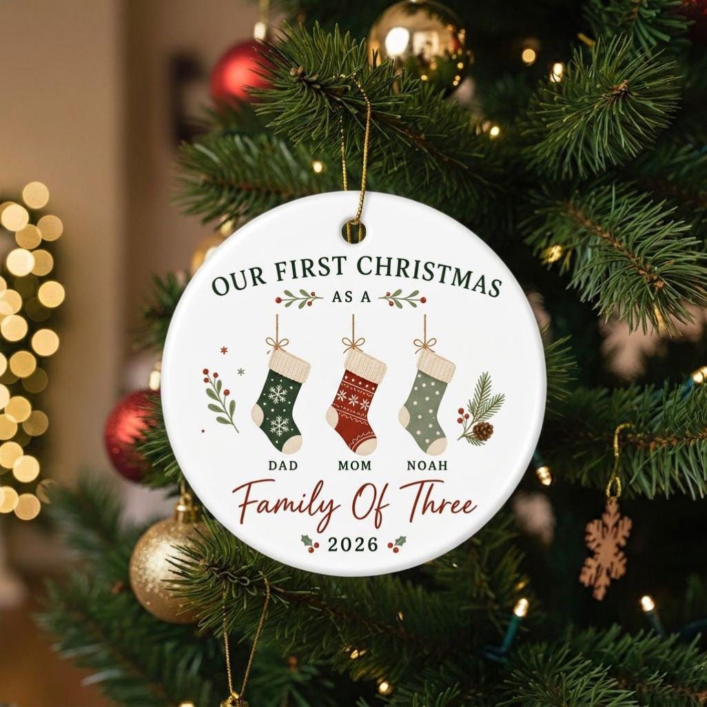 May include: A round white ceramic ornament featuring the text Our First Christmas As A Family Of Three 2026. The design includes three hanging stockings in green, red, and light green with patterns, labeled Dad, Mom, and Noah, with holly accents.