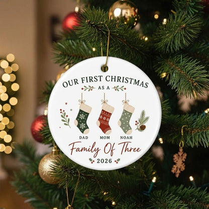 May include: A round white ceramic ornament featuring the text Our First Christmas As A Family Of Three 2026. The design includes three hanging stockings in green, red, and light green with patterns, labeled Dad, Mom, and Noah, with holly accents.