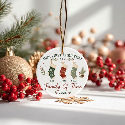 May include: A round white ceramic Christmas ornament hanging from a gold string. It features three illustrated stockings labeled Dad, Mom, and Noah, with the text Our First Christmas As A Family Of Three and the year 2026 printed in red and green.