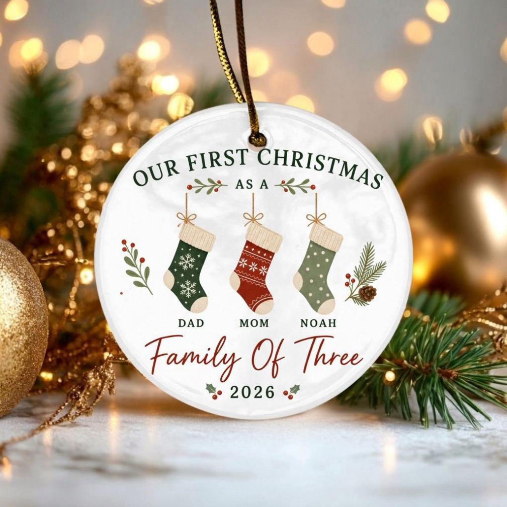 May include: A round white ceramic Christmas ornament hanging from a gold string. It features the text "OUR FIRST CHRISTMAS AS A Family Of Three" with three hanging stockings labeled "DAD", "MOM", and "NOAH" above the year "2026" in red and green.