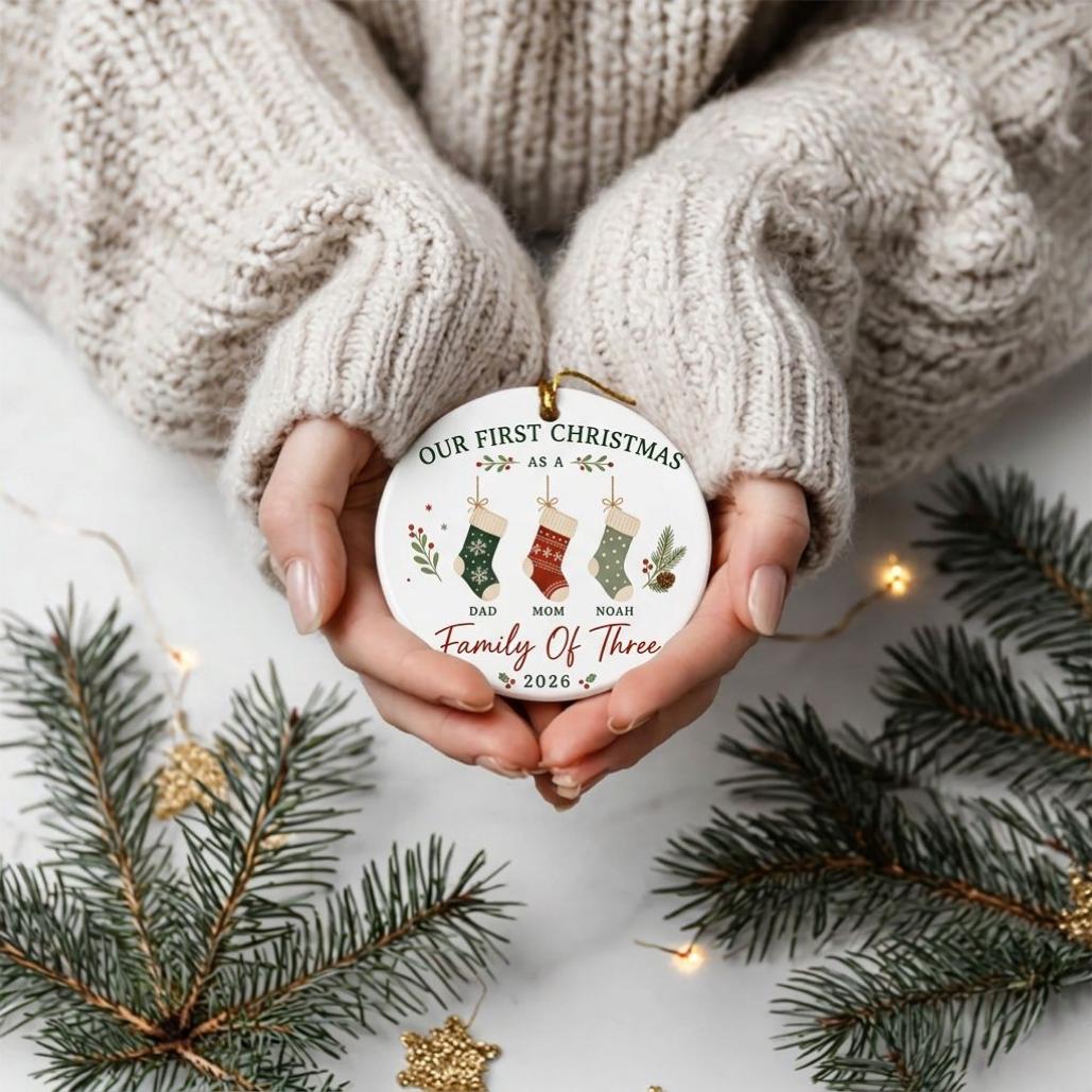 May include: A round white ceramic ornament featuring the text "OUR FIRST CHRISTMAS" and "Family Of Three 2026". The design includes three hanging stockings labeled "DAD", "MOM", and "NOAH" with green, red, and light green patterns and pine sprigs.