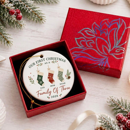 May include: A round white ceramic ornament featuring the text "OUR FIRST CHRISTMAS AS A Family Of Three 2026" with illustrations of three hanging stockings labeled "DAD", "MOM", and "NOAH". The ornament is displayed inside an open red gift box.
