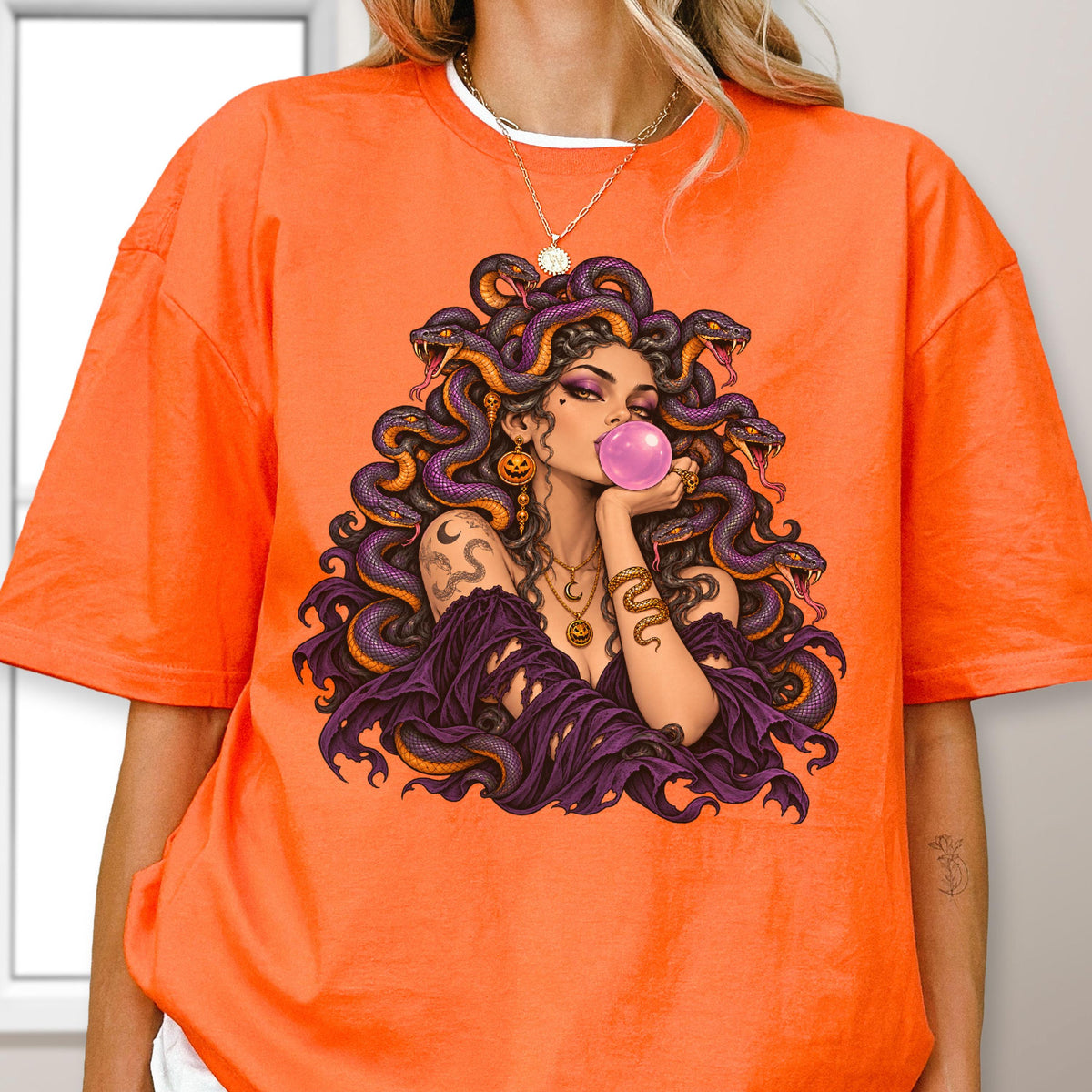 May include: An orange t-shirt featuring a graphic illustration of a figure with snakes for hair, blowing a pink bubblegum bubble. The figure wears purple clothing and gold jewelry, including a necklace with a crescent moon and pumpkin charm.