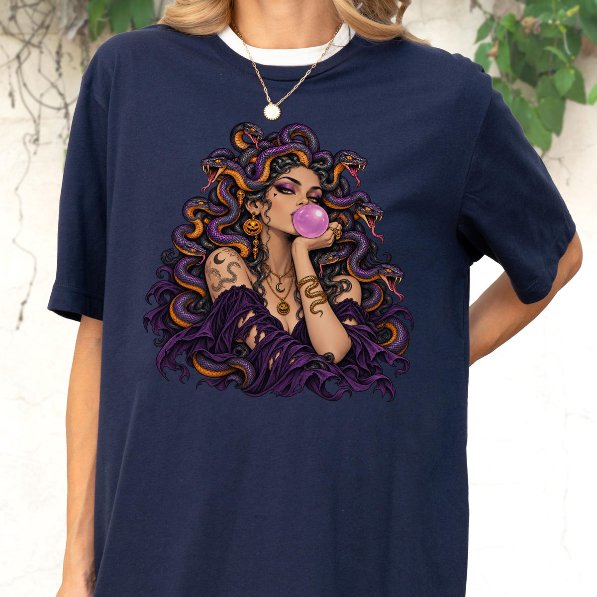 May include: A navy blue short-sleeve t-shirt featuring a graphic illustration of a figure with snakes for hair, wearing a purple garment, and blowing a pink bubble. The figure is adorned with gold jewelry, including a necklace and a snake arm cuff.