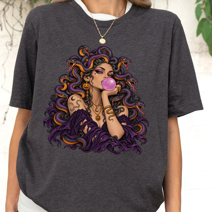 May include: A charcoal gray t-shirt featuring a graphic print of a figure with snakes for hair, wearing a purple garment, blowing a pink bubblegum bubble, and adorned with gold jewelry including pumpkin-shaped earrings and a crescent moon necklace.