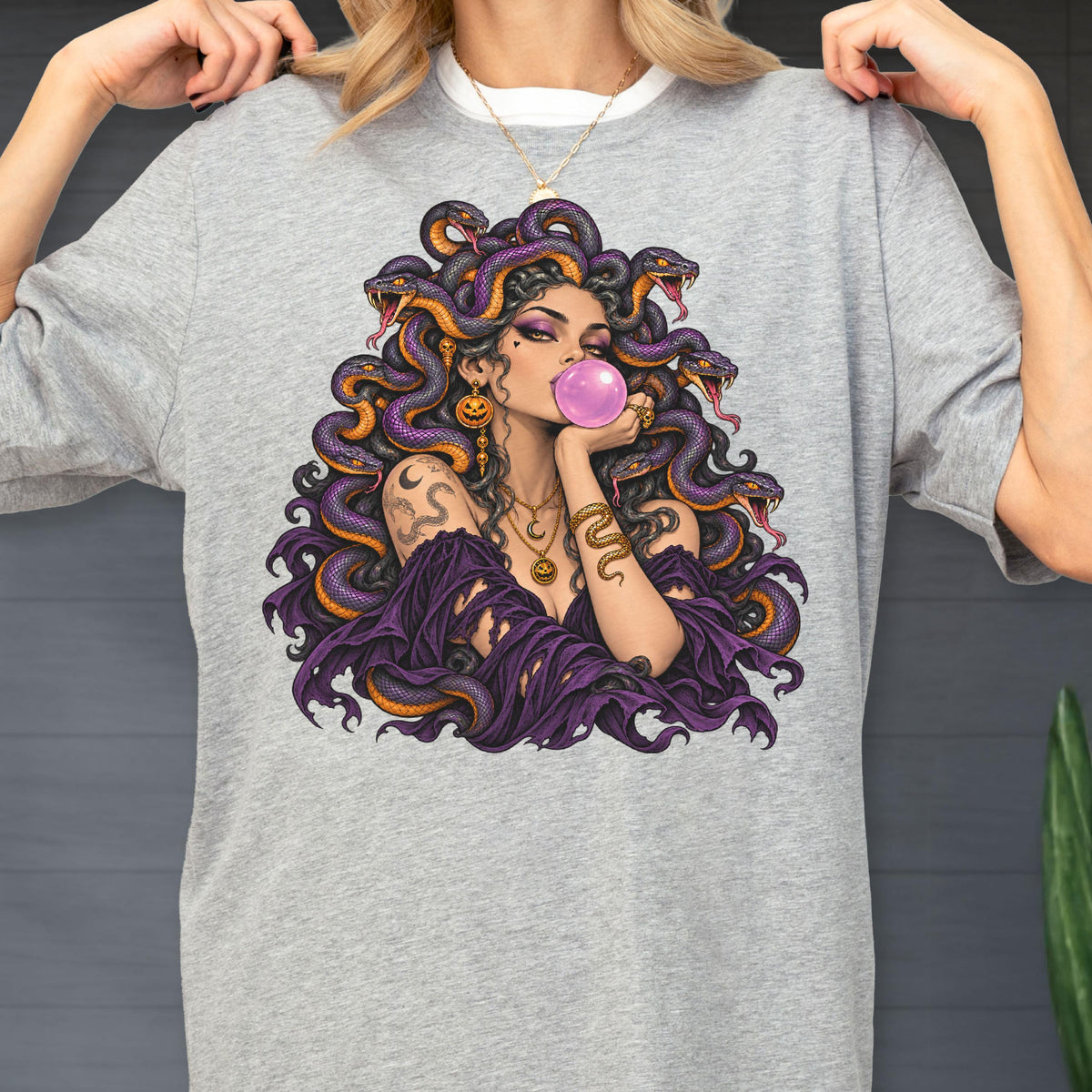 May include: A heather gray t-shirt featuring a graphic illustration of a figure with purple snakes for hair, blowing a pink bubblegum bubble. The figure wears a dark purple off-the-shoulder garment, gold necklaces, and snake-themed arm tattoos.