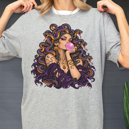 May include: A heather gray t-shirt featuring a graphic illustration of a figure with purple snakes for hair, blowing a pink bubblegum bubble. The figure wears a dark purple off-the-shoulder garment, gold necklaces, and snake-themed arm tattoos.
