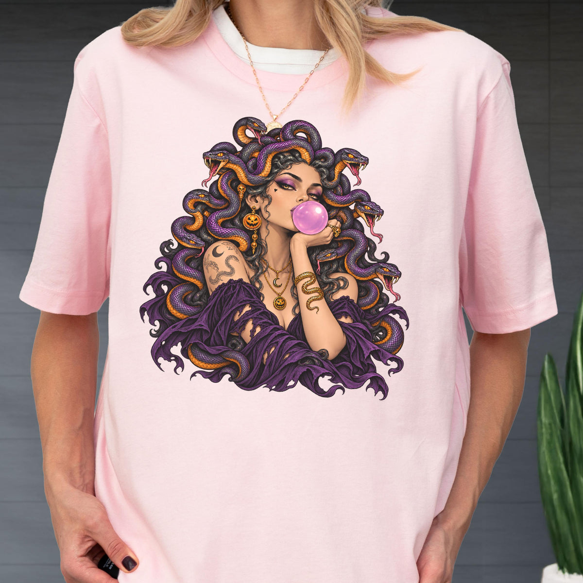May include: A light pink t-shirt featuring a graphic illustration of a figure with purple snakes for hair, blowing a pink bubblegum bubble. The figure wears a dark purple garment, gold necklaces, and gold earrings, with a snake tattoo on the arm.