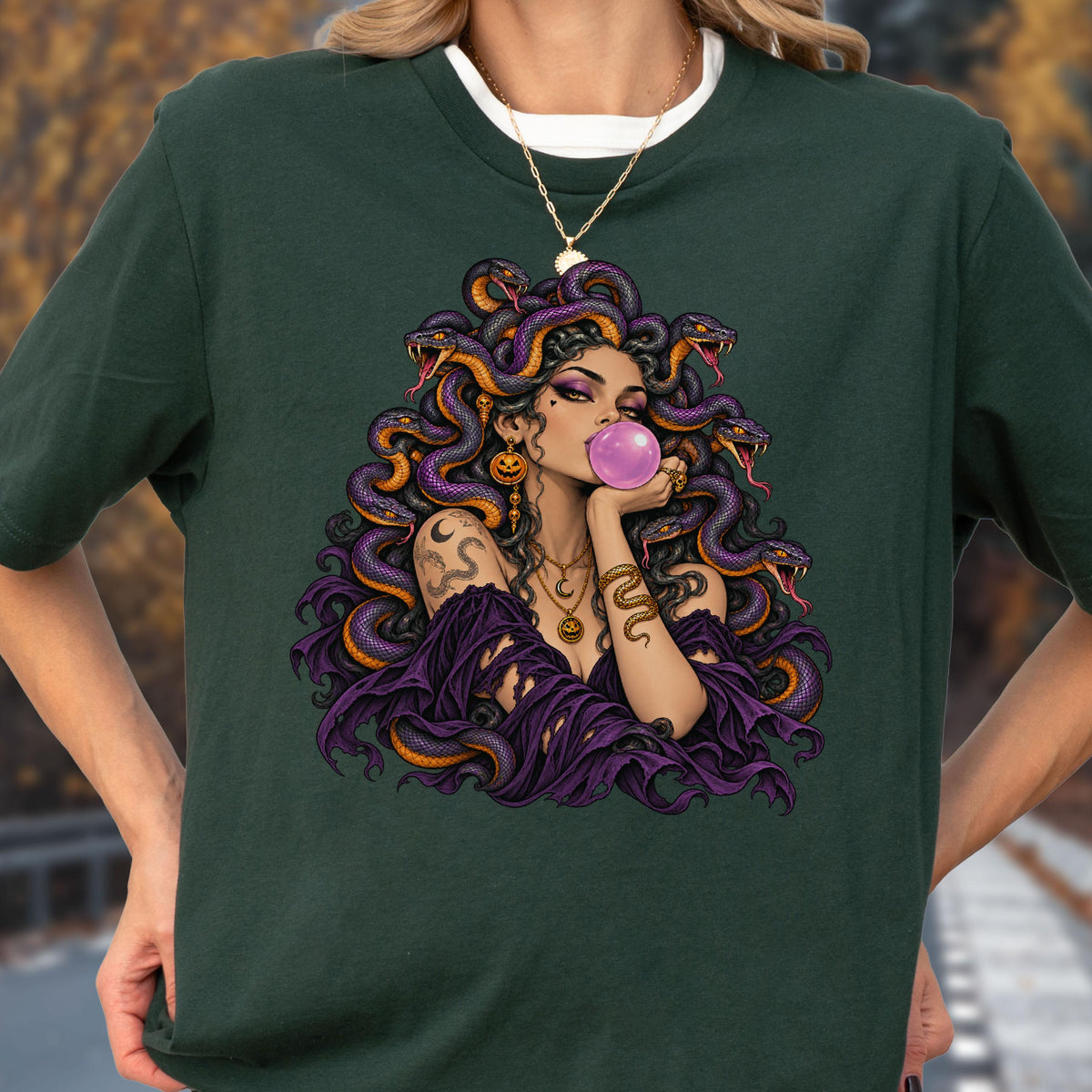 May include: A dark green t-shirt featuring a graphic illustration of a figure with purple snakes for hair, blowing a pink bubblegum bubble. The figure wears a purple off-the-shoulder top, layered gold necklaces, and a snake arm cuff.