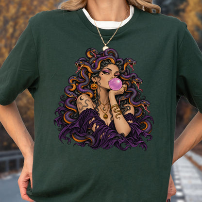 May include: A dark green t-shirt featuring a graphic illustration of a figure with purple snakes for hair, blowing a pink bubblegum bubble. The figure wears a purple off-the-shoulder top, layered gold necklaces, and a snake arm cuff.