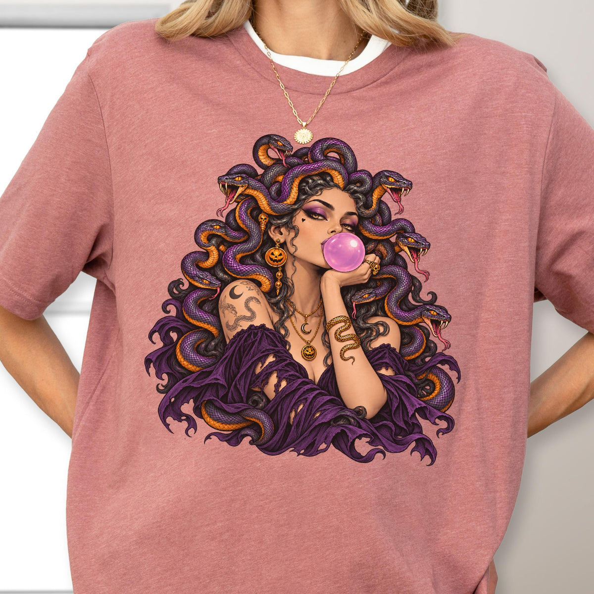 May include: A dusty rose t-shirt featuring a graphic illustration of a figure with snakes for hair, blowing a pink bubblegum bubble. The figure wears layered gold necklaces and has a snake tattoo on the arm, set against a purple draped garment.