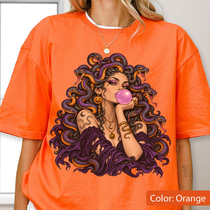 May include: An orange t-shirt featuring a graphic illustration of a figure with purple snakes for hair, blowing a pink bubblegum bubble. The figure wears layered gold necklaces and has a snake tattoo on the arm, set against the bright orange fabric.
