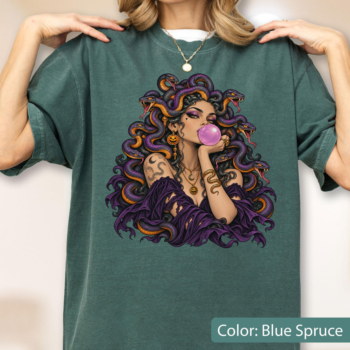 May include: A blue spruce t-shirt featuring a graphic illustration of a figure with purple snakes for hair, blowing a pink bubblegum bubble. The figure wears layered gold necklaces and a purple garment, surrounded by multiple detailed snakes.