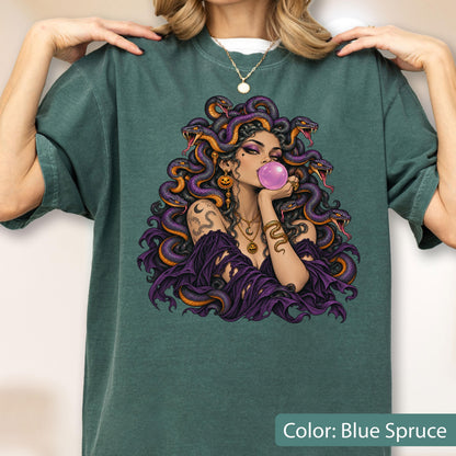 May include: A blue spruce t-shirt featuring a graphic illustration of a figure with purple snakes for hair, blowing a pink bubblegum bubble. The figure wears layered gold necklaces and a purple garment, surrounded by multiple detailed snakes.