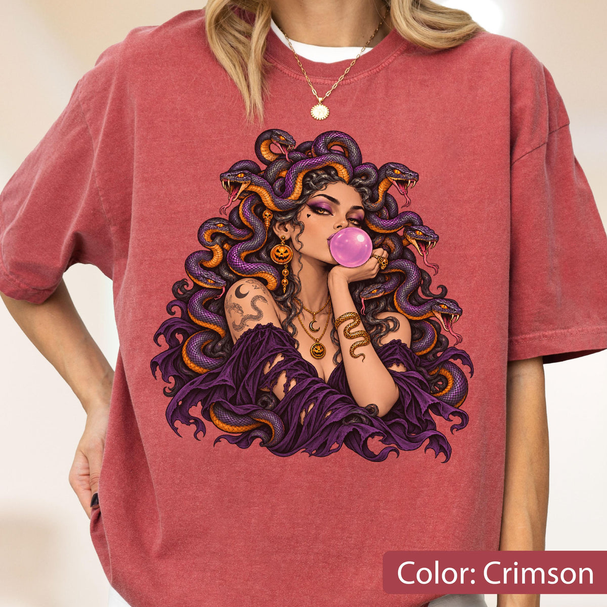 May include: A crimson t-shirt featuring a graphic print of a figure with purple snakes for hair, blowing a pink bubblegum bubble. The figure wears a purple off-the-shoulder top, layered gold necklaces, and a snake-shaped arm cuff.