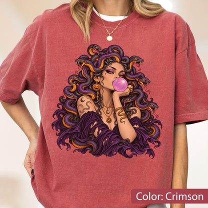 May include: A crimson t-shirt featuring a graphic print of a figure with purple snakes for hair, blowing a pink bubblegum bubble. The figure wears a purple off-the-shoulder top, layered gold necklaces, and a snake-shaped arm cuff.