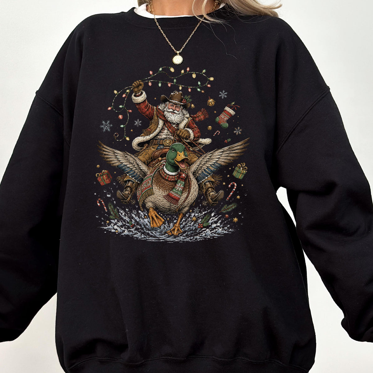 May include: A black crewneck sweatshirt featuring a graphic print of Santa Claus wearing a cowboy hat and vest, riding a flying mallard duck. The design includes colorful Christmas lights, snowflakes, gift boxes, and candy canes in the background.