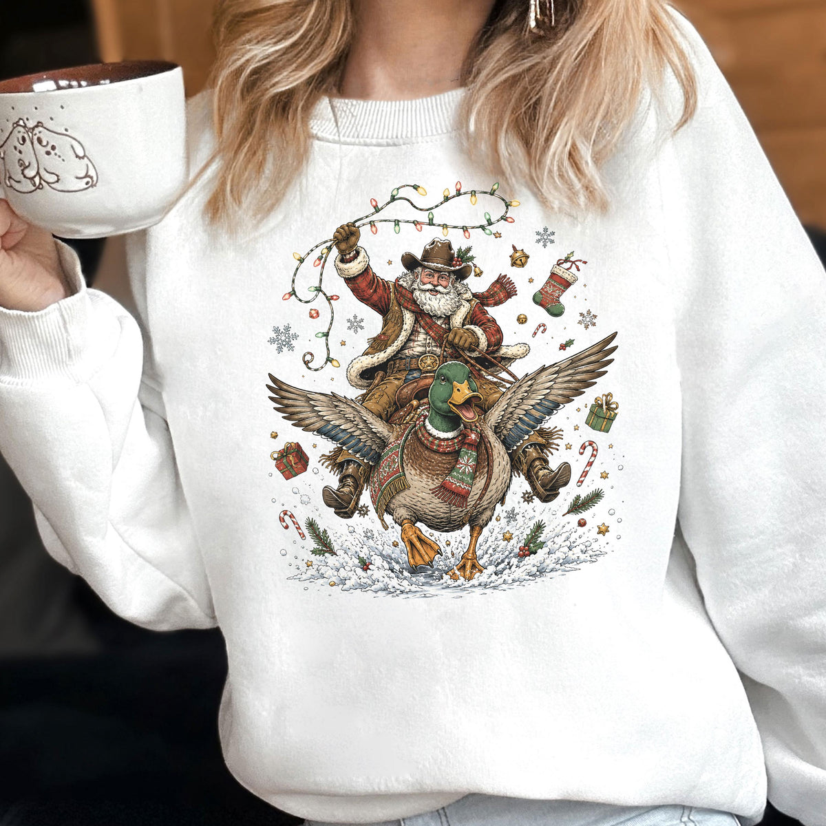 May include: A white crewneck sweatshirt featuring a graphic print of a cowboy Santa Claus riding a flying mallard duck. The design includes colorful Christmas lights, snowflakes, candy canes, and gift boxes surrounding the central figures.