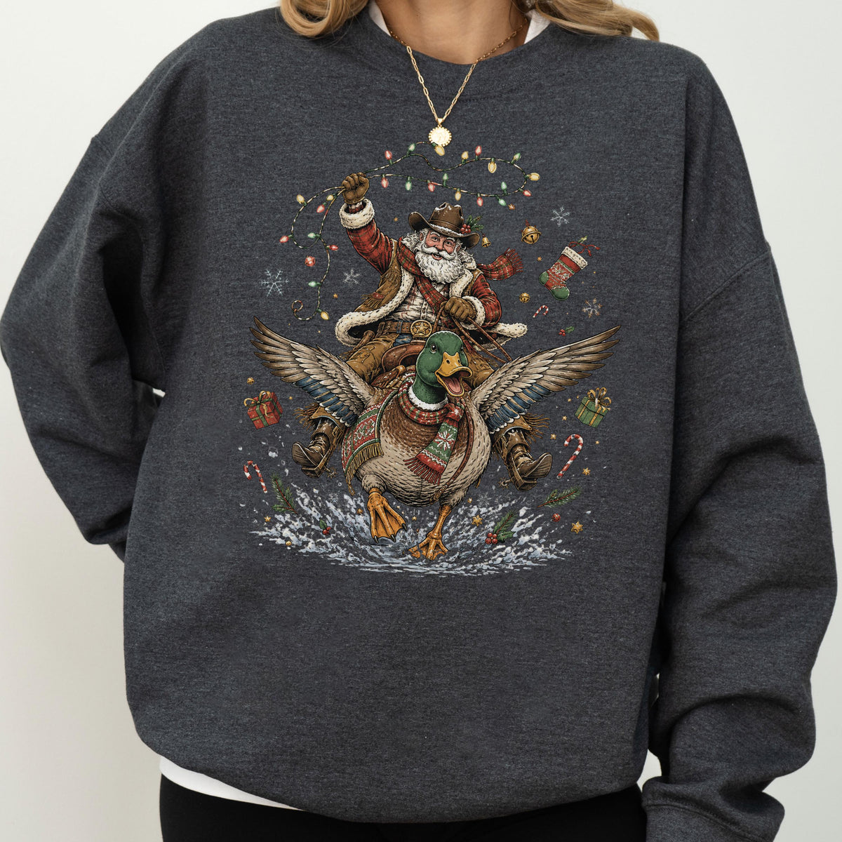 May include: A dark charcoal gray crewneck sweatshirt featuring a graphic print of Santa Claus wearing a cowboy hat and vest, riding a flying mallard duck. The design includes colorful Christmas lights, snowflakes, gift boxes, and candy canes.