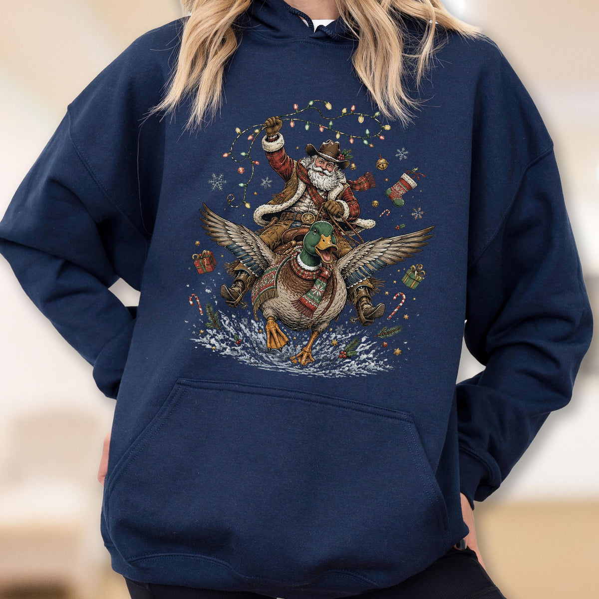 May include: A navy blue hooded sweatshirt featuring a graphic print of Santa Claus wearing a cowboy hat and riding a flying mallard duck. The design includes Christmas lights, snowflakes, gift boxes, a stocking, and candy canes surrounding them.