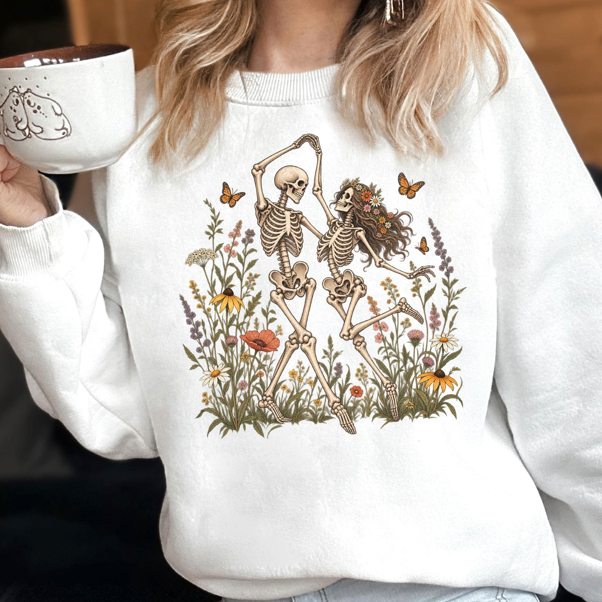 May include: A white crewneck sweatshirt featuring a graphic print of two skeletons dancing amidst a colorful field of wildflowers and butterflies. The skeletons are depicted in a joyful pose, surrounded by various botanical elements and orange butterflies.