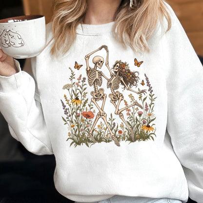 May include: A white crewneck sweatshirt featuring a graphic print of two skeletons dancing amidst a colorful field of wildflowers and butterflies. The skeletons are depicted in a joyful pose, surrounded by various botanical elements and orange butterflies.