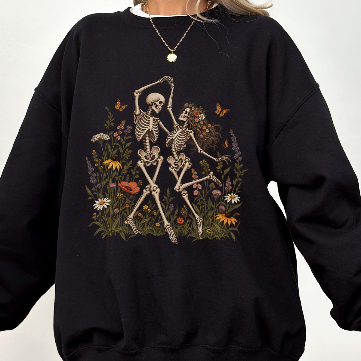 May include: A black crewneck sweatshirt featuring a graphic print of two skeletons dancing in a field of colorful wildflowers. The skeletons are surrounded by various blooming flowers, green foliage, and small orange butterflies in a detailed illustration.