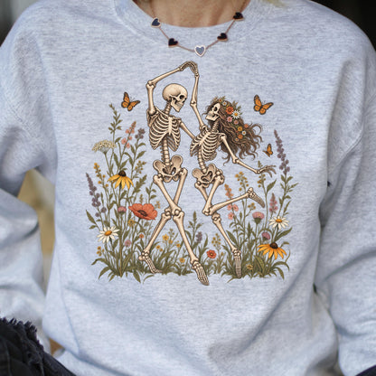 May include: A light gray crewneck sweatshirt featuring a graphic print of two skeletons dancing in a field of colorful wildflowers and orange butterflies. The skeleton on the right wears a crown of flowers in its long, flowing brown hair.