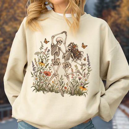 May include: A beige crewneck sweatshirt featuring a graphic print of two skeletons dancing amidst a field of colorful wildflowers and butterflies. One skeleton has long hair adorned with a floral crown, while the other is bald.