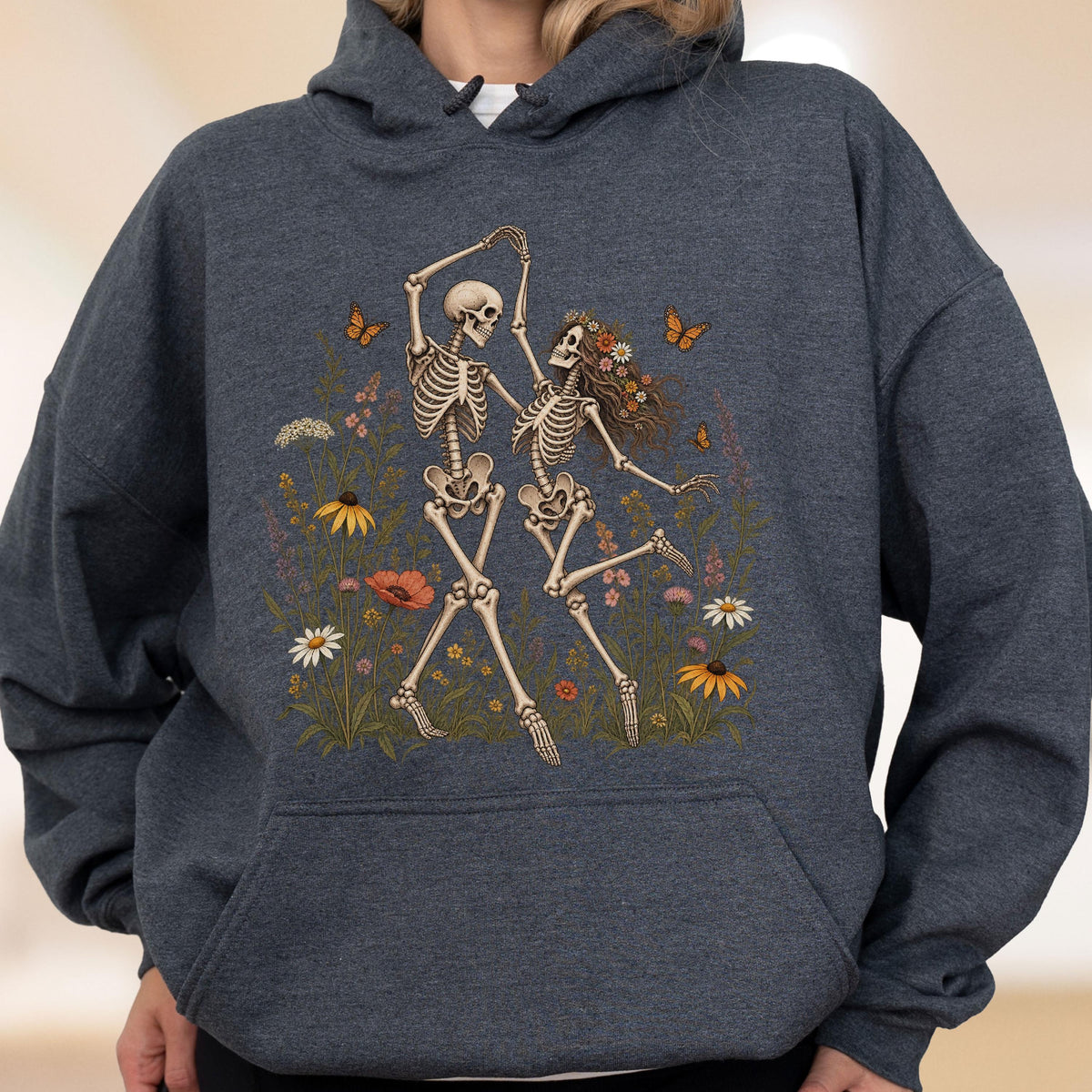 May include: A dark heather gray hooded sweatshirt featuring a graphic print of two skeletons dancing in a field of colorful wildflowers and orange butterflies. The hoodie includes a front kangaroo pocket, long sleeves, and a drawstring hood.