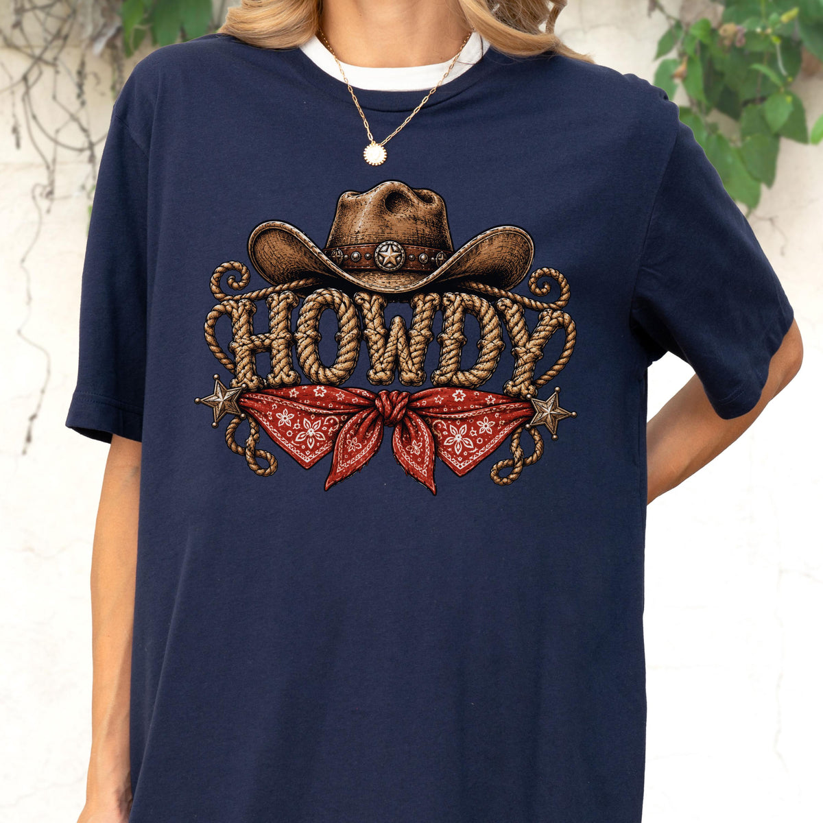 May include: A navy blue t-shirt featuring a graphic print of a brown cowboy hat above the word HOWDY written in a rope-style font. Below the text is a red bandana with a white floral pattern, flanked by two small metallic-colored stars.