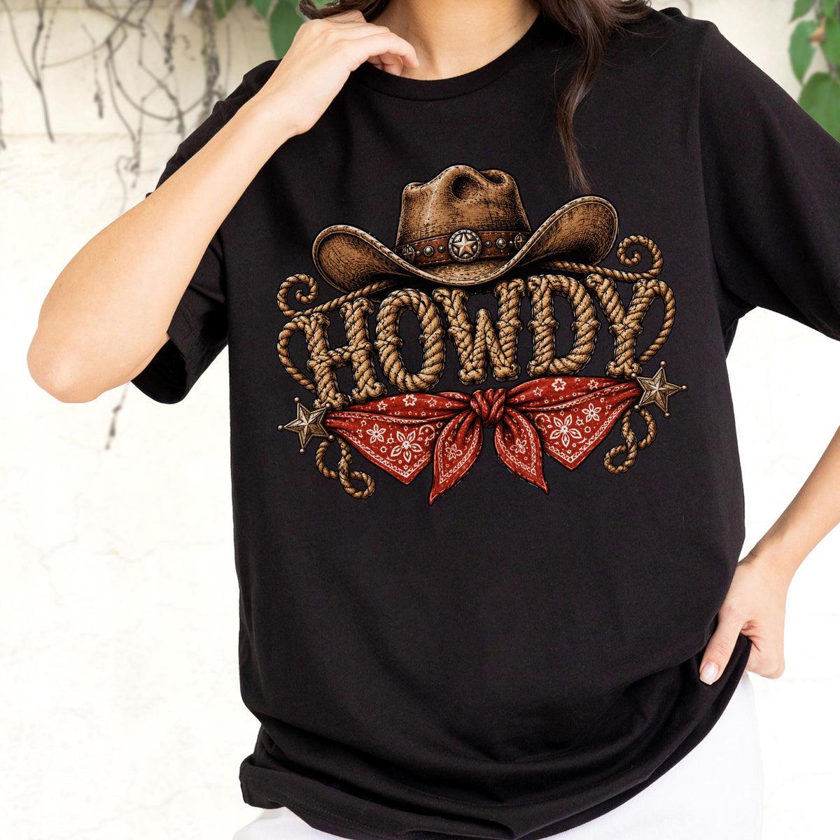 May include: A black t-shirt featuring a graphic print of a brown cowboy hat above the word HOWDY written in a rope-style font. Below the text is a red bandana with a white floral pattern, flanked by two small metallic-colored star icons.