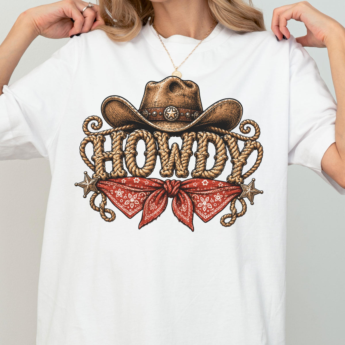 May include: A white t-shirt featuring a graphic print of a brown cowboy hat resting above the word HOWDY, which is written in a rope-textured font. Below the text is a red bandana with a white floral pattern, flanked by two silver stars.