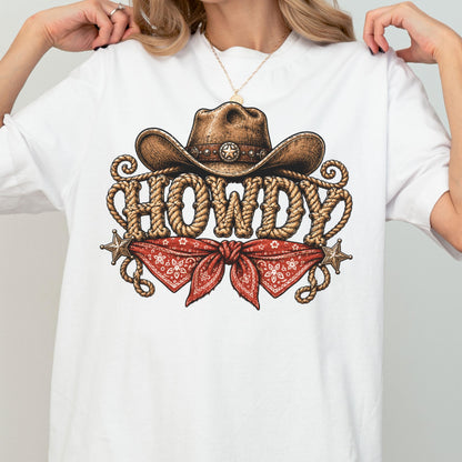 May include: A white t-shirt featuring a graphic print of a brown cowboy hat resting above the word HOWDY, which is written in a rope-textured font. Below the text is a red bandana with a white floral pattern, flanked by two silver stars.