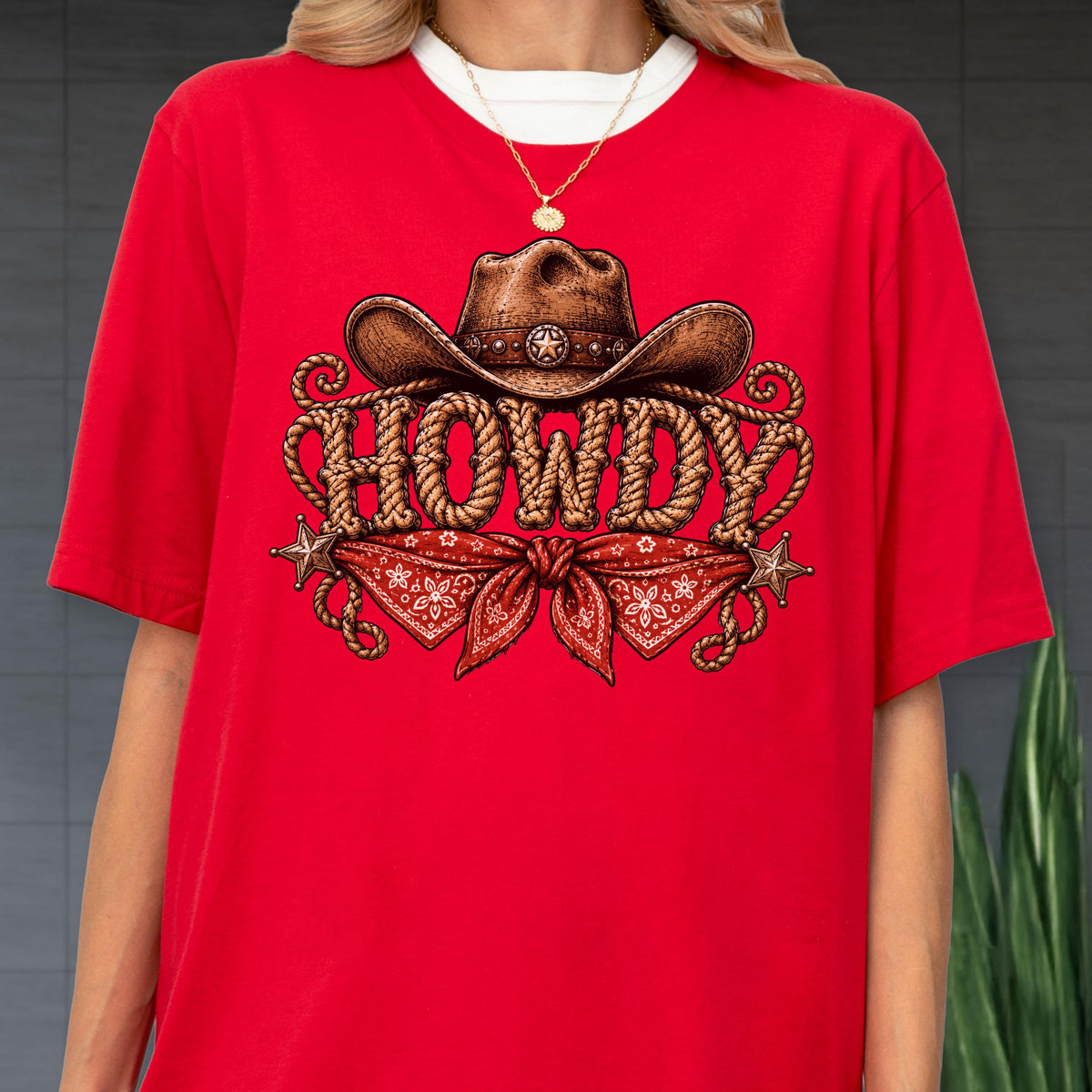 May include: A bright red short-sleeved t-shirt featuring a graphic print of a brown cowboy hat resting above the word HOWDY, which is written in a rope-style font. Below the text is a red bandana with a white floral pattern and two star icons.