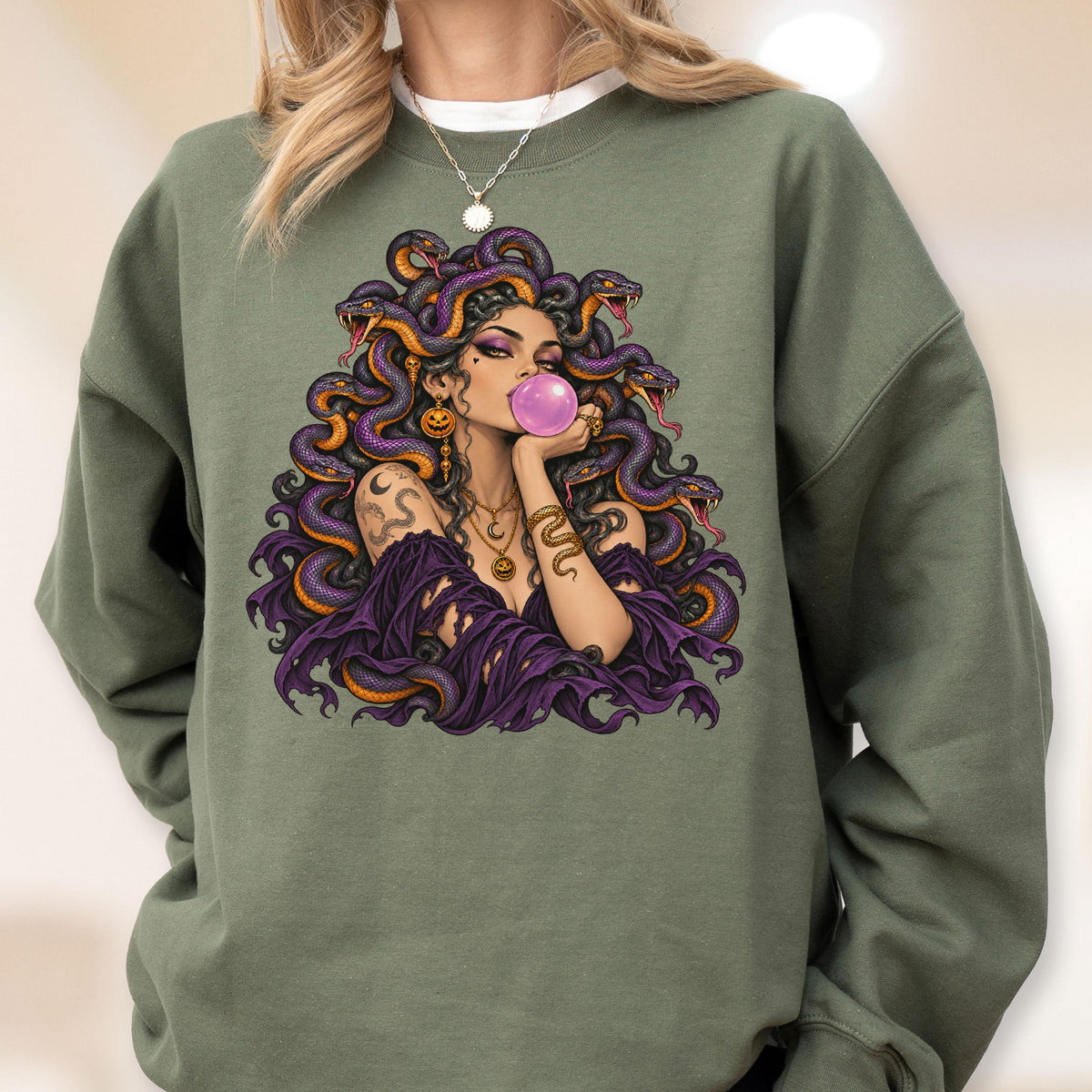 May include: An olive green crewneck sweatshirt featuring a graphic print of a figure with purple snakes for hair, blowing a pink bubblegum bubble. The figure wears a dark purple off-the-shoulder top, gold jewelry, and has visible tattoos.