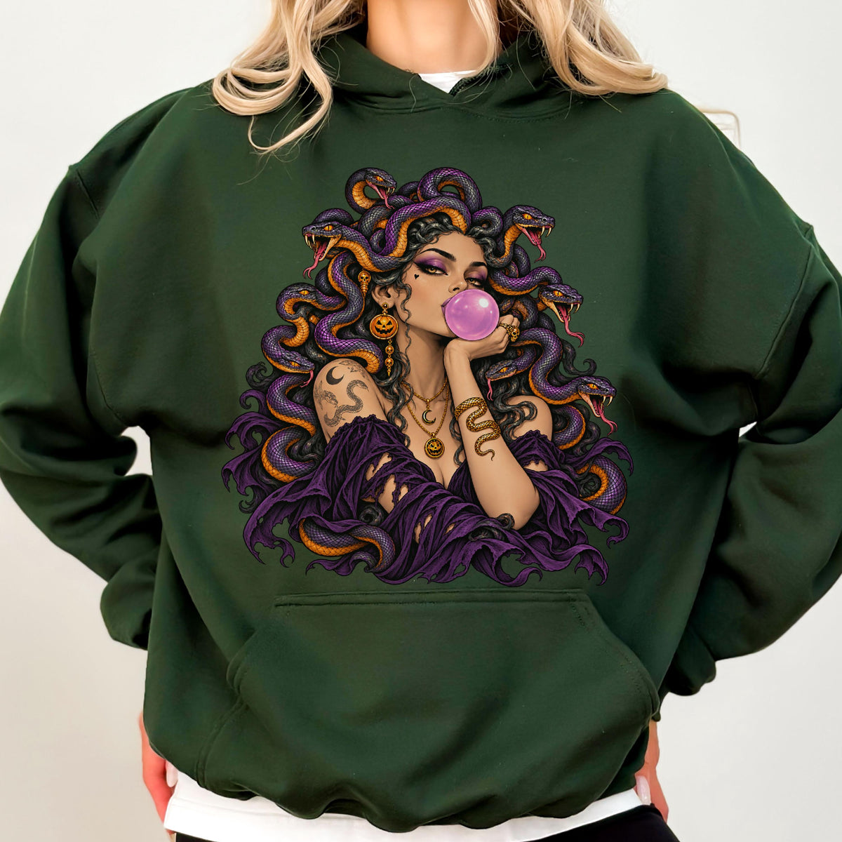 May include: A dark forest green hooded sweatshirt featuring a graphic illustration of a figure with snakes for hair, blowing a pink bubble. The figure wears layered necklaces and a dark purple garment, surrounded by purple and orange snakes.