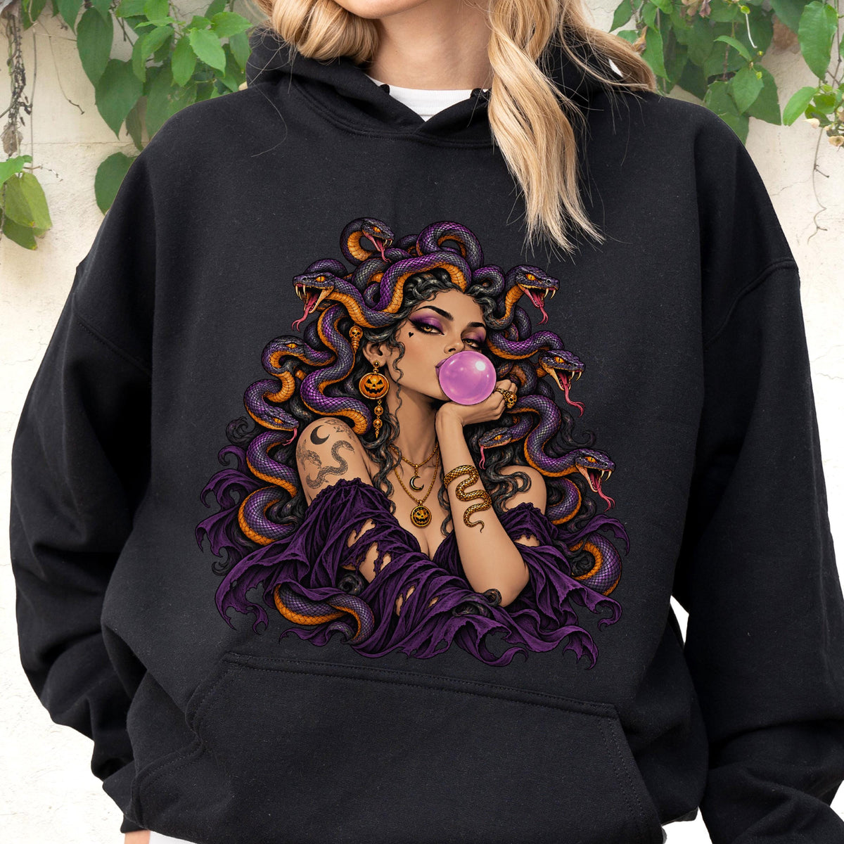 May include: A black hooded sweatshirt featuring a graphic print of a figure with purple snakes for hair, blowing a pink bubble. The figure wears a dark purple off-the-shoulder garment, gold necklaces with pumpkin charms, and has a snake tattoo on the arm.