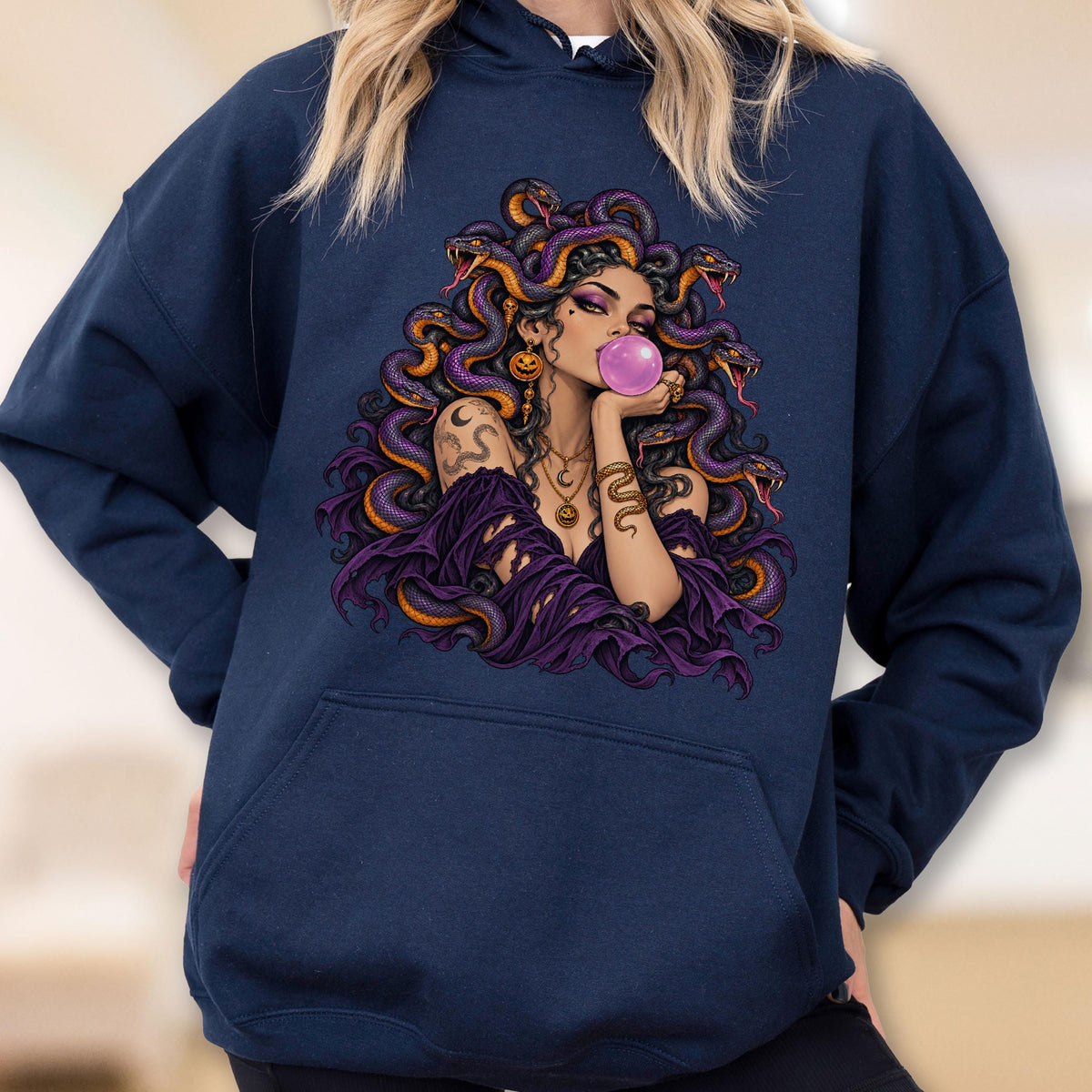 May include: A navy blue hooded sweatshirt featuring a graphic print of a figure with snakes for hair, wearing a purple off-the-shoulder top and gold jewelry, blowing a pink bubble. The design includes multiple snakes with orange and purple scales.