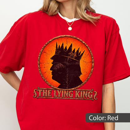 May include: A bright red short-sleeved t-shirt featuring a circular graphic with a cracked orange background. Inside the circle is a dark silhouette of a person wearing a crown. Below the graphic, the text reads THE LYING KING in a bold font.