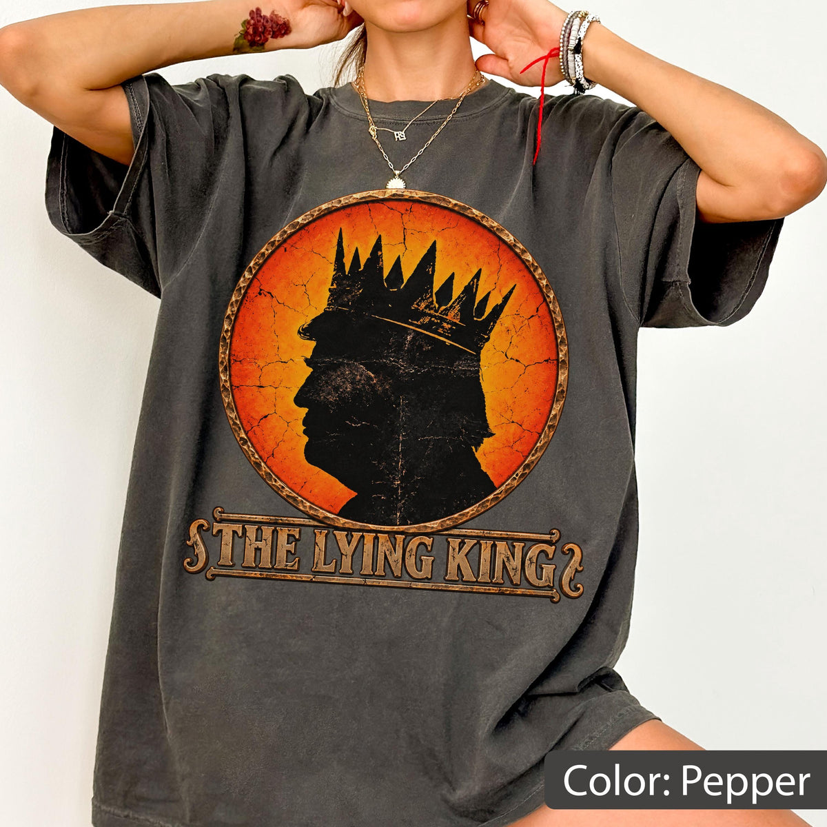 May include: A pepper-colored short-sleeve t-shirt featuring a circular graphic with a distressed orange background. The graphic depicts a black silhouette of a person wearing a crown, with the text "THE LYING KING" printed in a bold, serif font below.