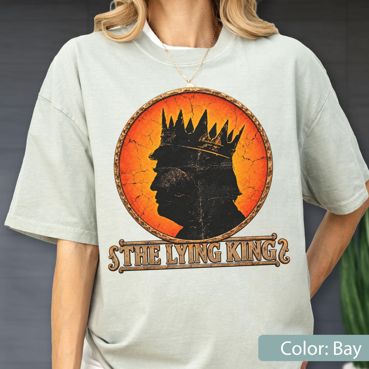 May include: A bay-colored t-shirt featuring a circular graphic with a distressed orange background. Inside the circle is a black silhouette of a person wearing a crown. Below the graphic, the text reads THE LYING KING in a stylized font.