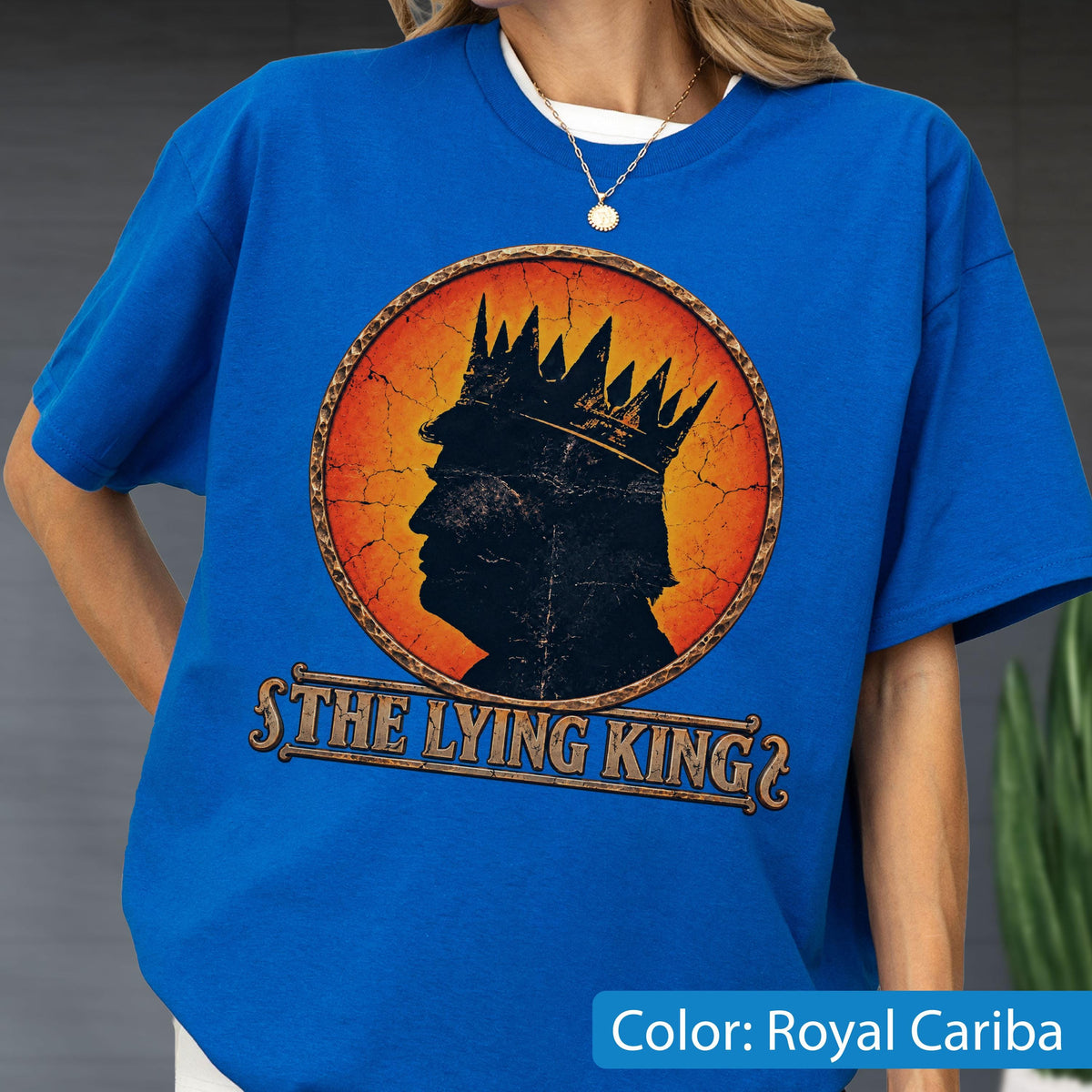 May include: A royal blue t-shirt featuring a circular graphic with a distressed orange and yellow background. The graphic depicts a black silhouette of a person wearing a crown, with the text "THE LYING KING" printed in a bold, serif font below.